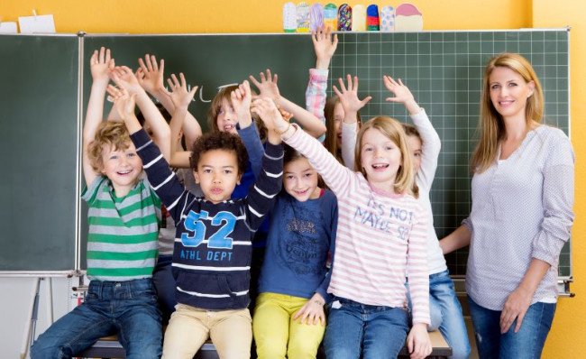 Maestra de preescolar y alumnos con las manos en alto frente a la pizarra